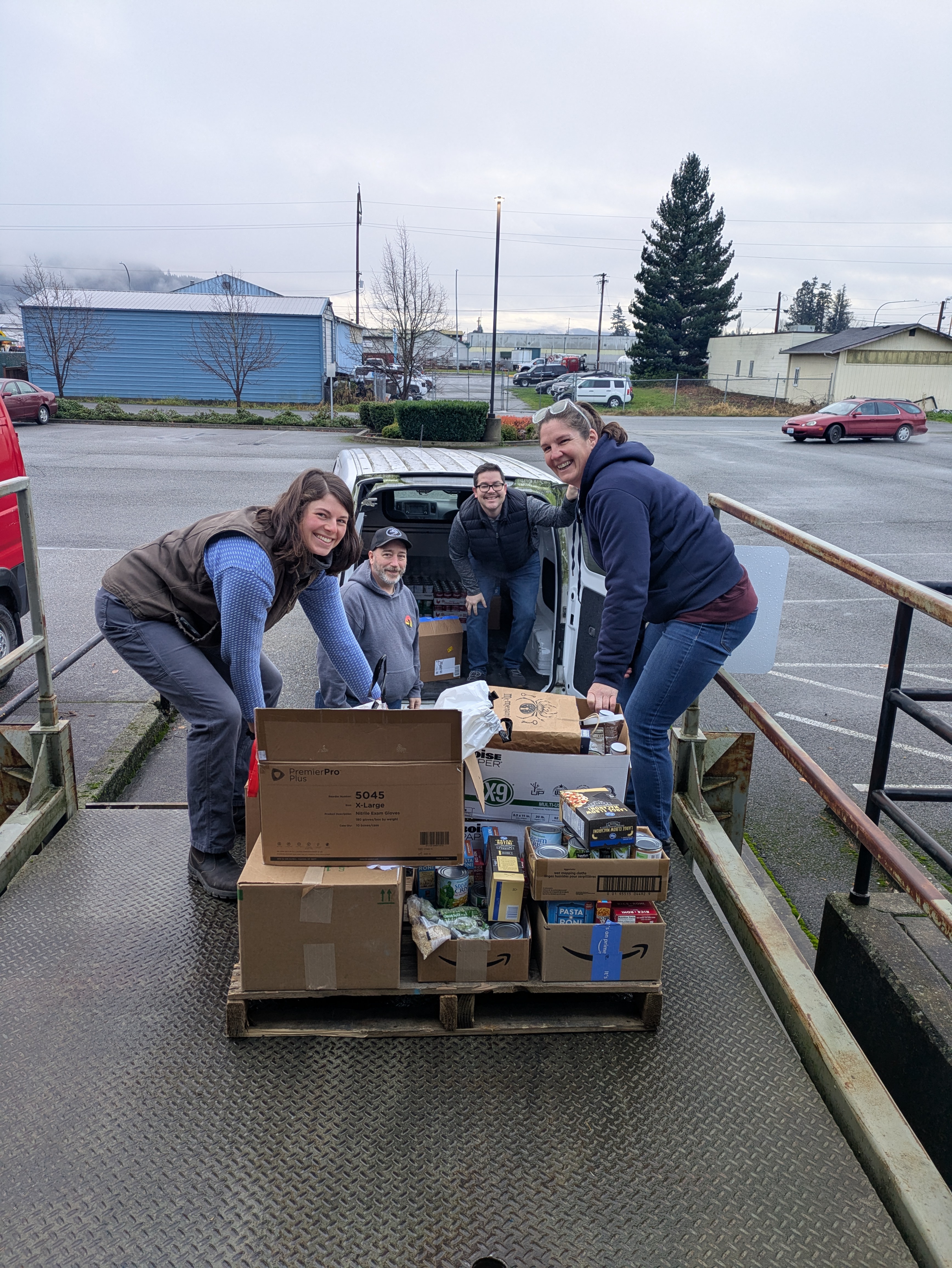 Two women and two men unload boxes of boxed and canned food from a white van.