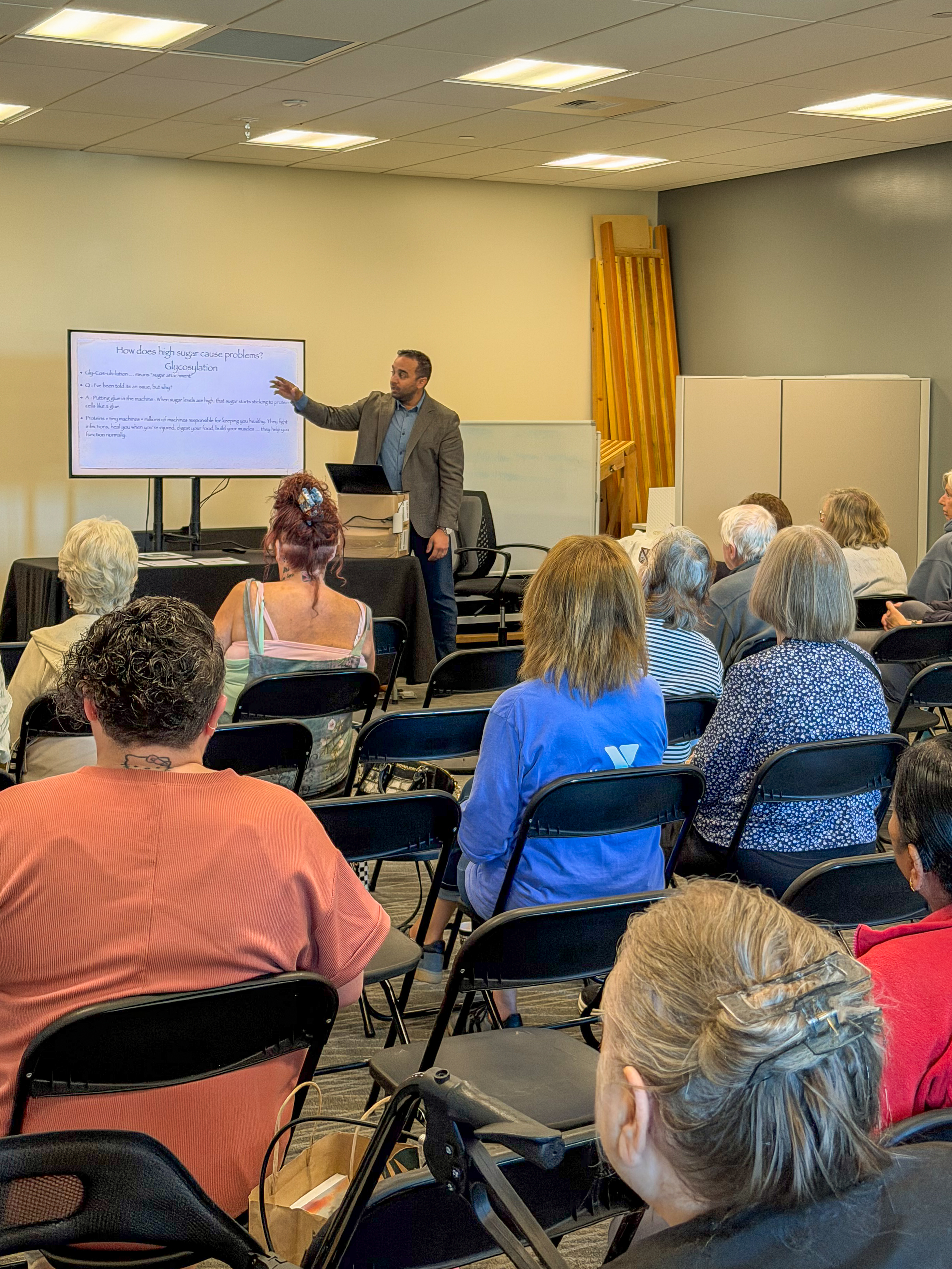 A presenter stands at the front of a meeting room, pointing to a slide displayed on a large monitor while speaking to an audience seated in rows of black folding chairs. 