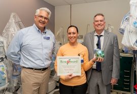 Un hombre, una mujer y otro hombre posan en una foto. Una mujer sistiene el certificado del premio Rose Award.