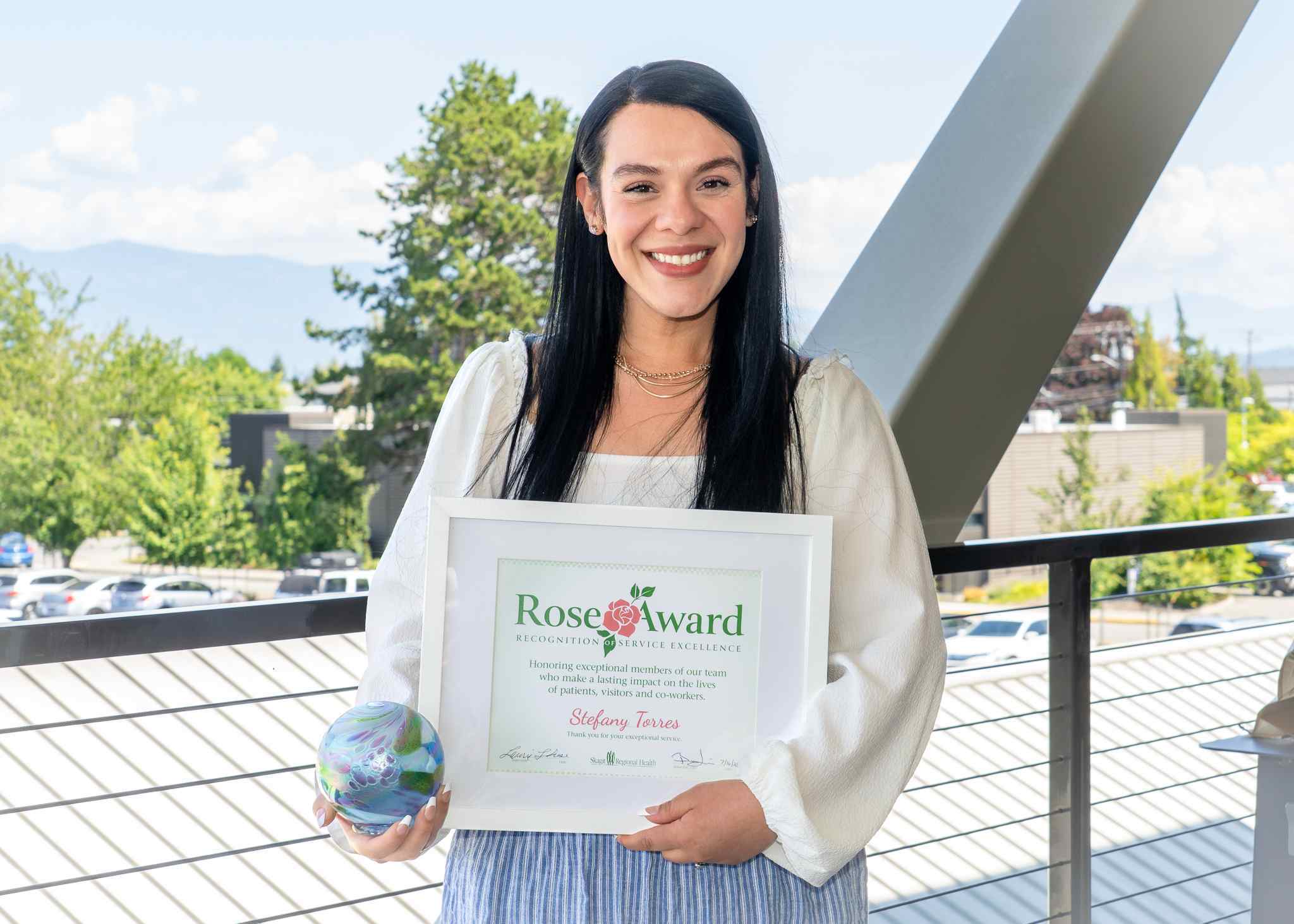 A woman standing on a balcony on a sunny day with an award she received.
