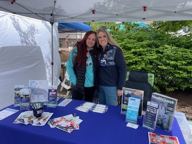 Two women stand at a table set-up at an outdoor health fair