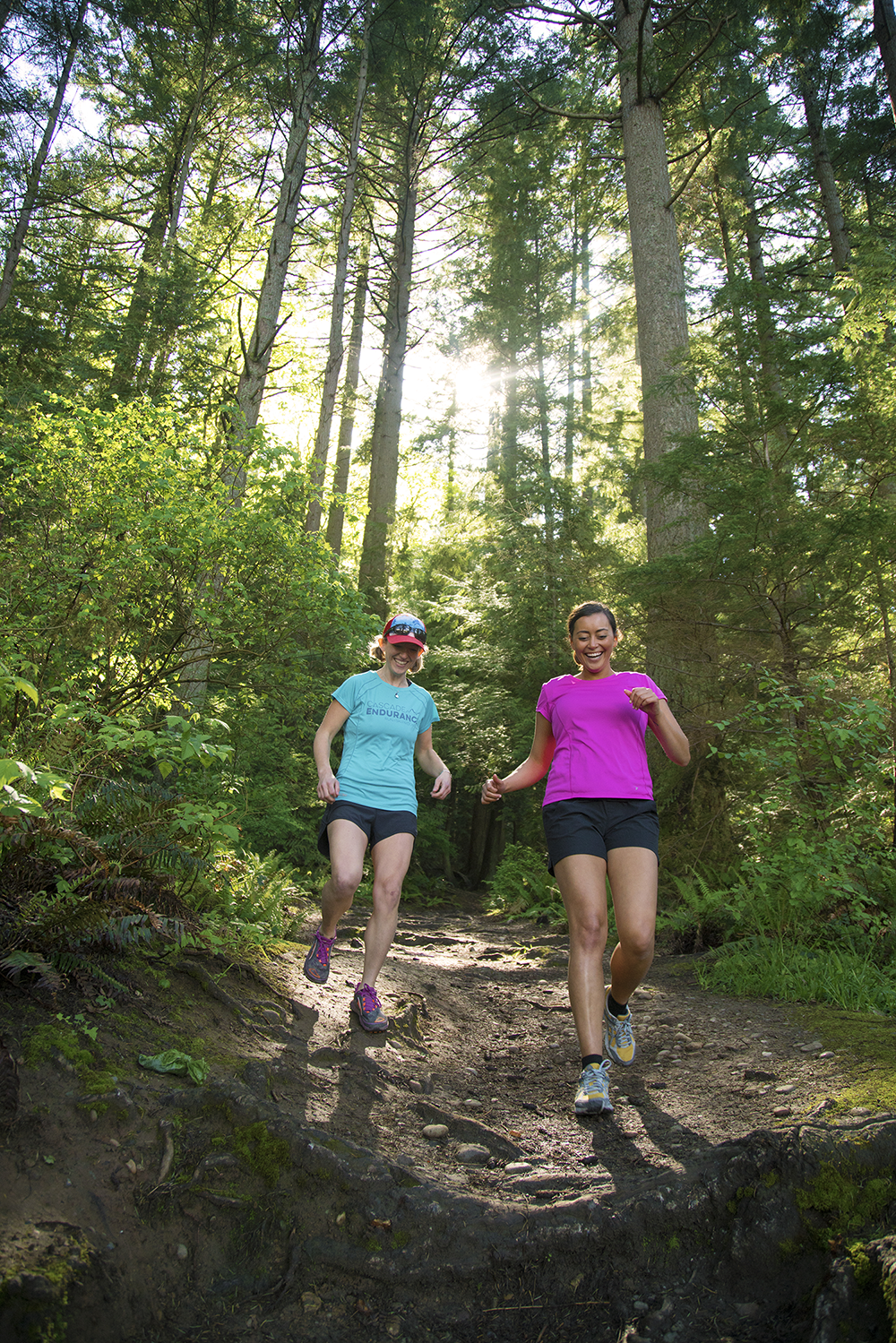 Two women run down a trail with a forest shown behind them. 